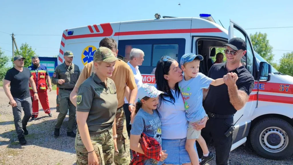 Жінка цілує хлопчика біля карети швидкої, поруч діти та військові