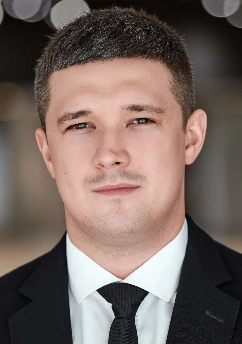 Чоловік у чорному костюмі з білою сорочкою та чорною краваткою дивиться в камеру