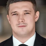 Чоловік у чорному костюмі з білою сорочкою та чорною краваткою дивиться в камеру