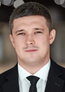 Чоловік у чорному костюмі з білою сорочкою та чорною краваткою дивиться в камеру