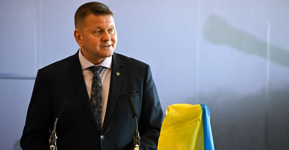 Чоловік у костюмі виступає за трибуною з мікрофонами біля українського прапора