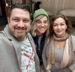 Троє людей усміхаються на селфі в приміщенні: бородатий чоловік, жінка в зеленій шапці та жінка з шарфом